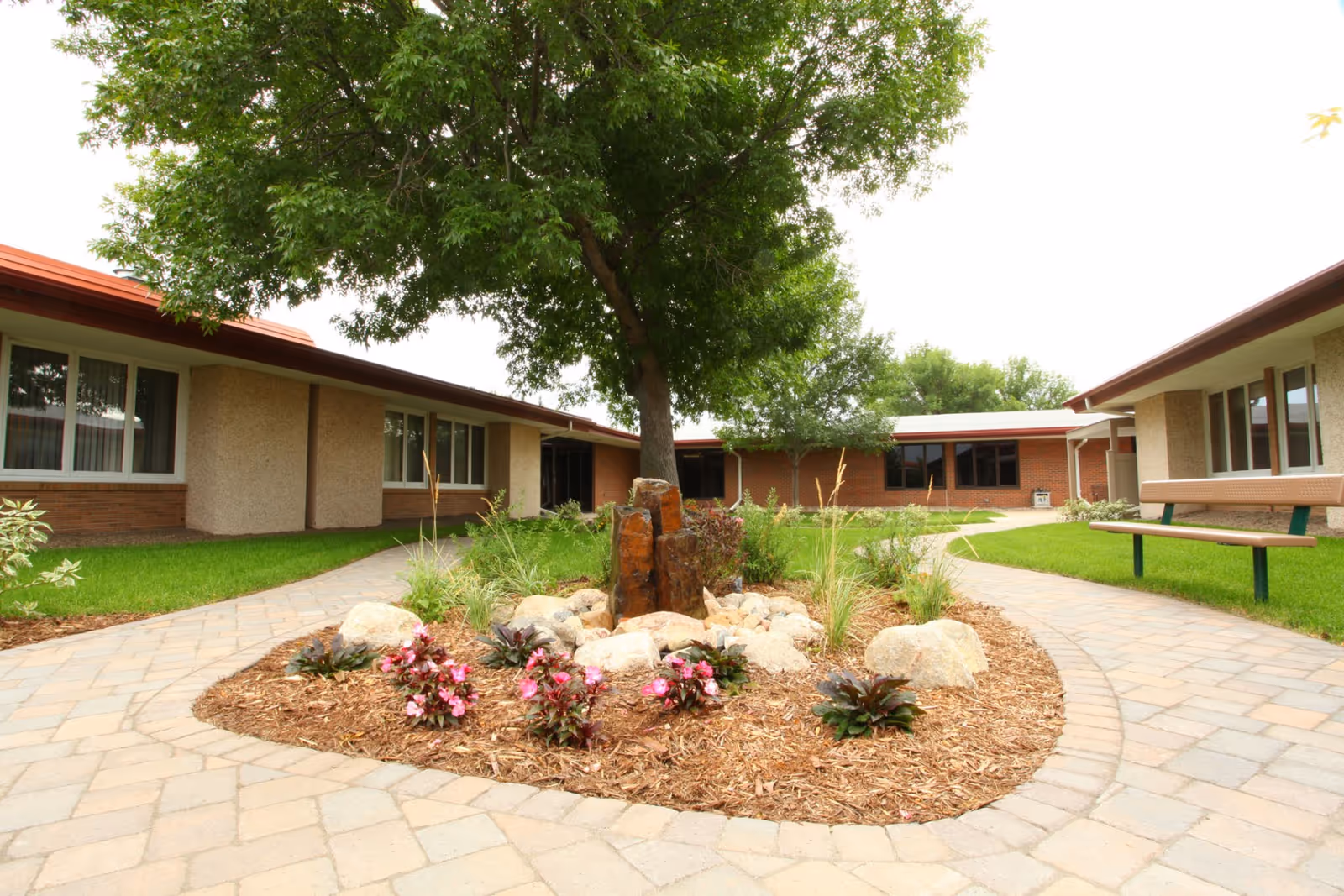 Courtyard with paved walkways, a central landscaped bed and tree, benches, and surrounding single-story brick buildings.