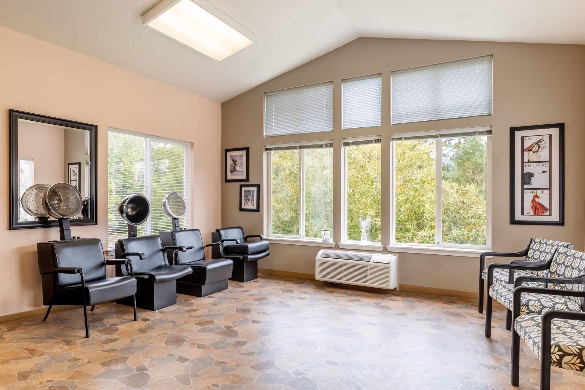 A bright and spacious hair salon area in a senior living community with three black salon chairs equipped with hair dryers on the left side, a large mirror on the wall, and three patterned chairs on the right side. Large windows with blinds let in natural light and show green trees outside.
