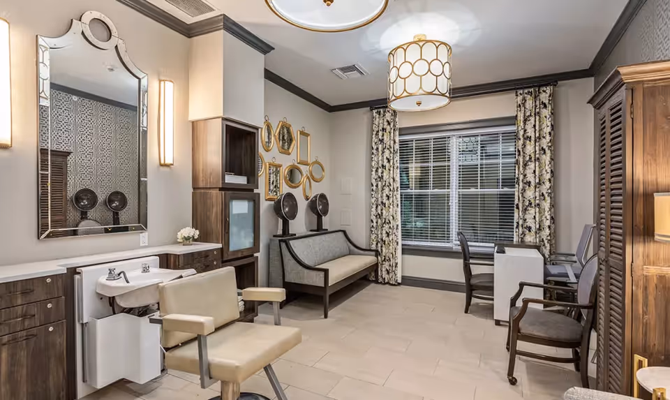 A well-lit interior room with a salon chair and sink on the left, a cushioned bench against the wall with decorative mirrors above it, a window with floral curtains, and a small desk with chairs near the window. The room has light-colored tiled flooring and neutral-colored walls with dark trim.