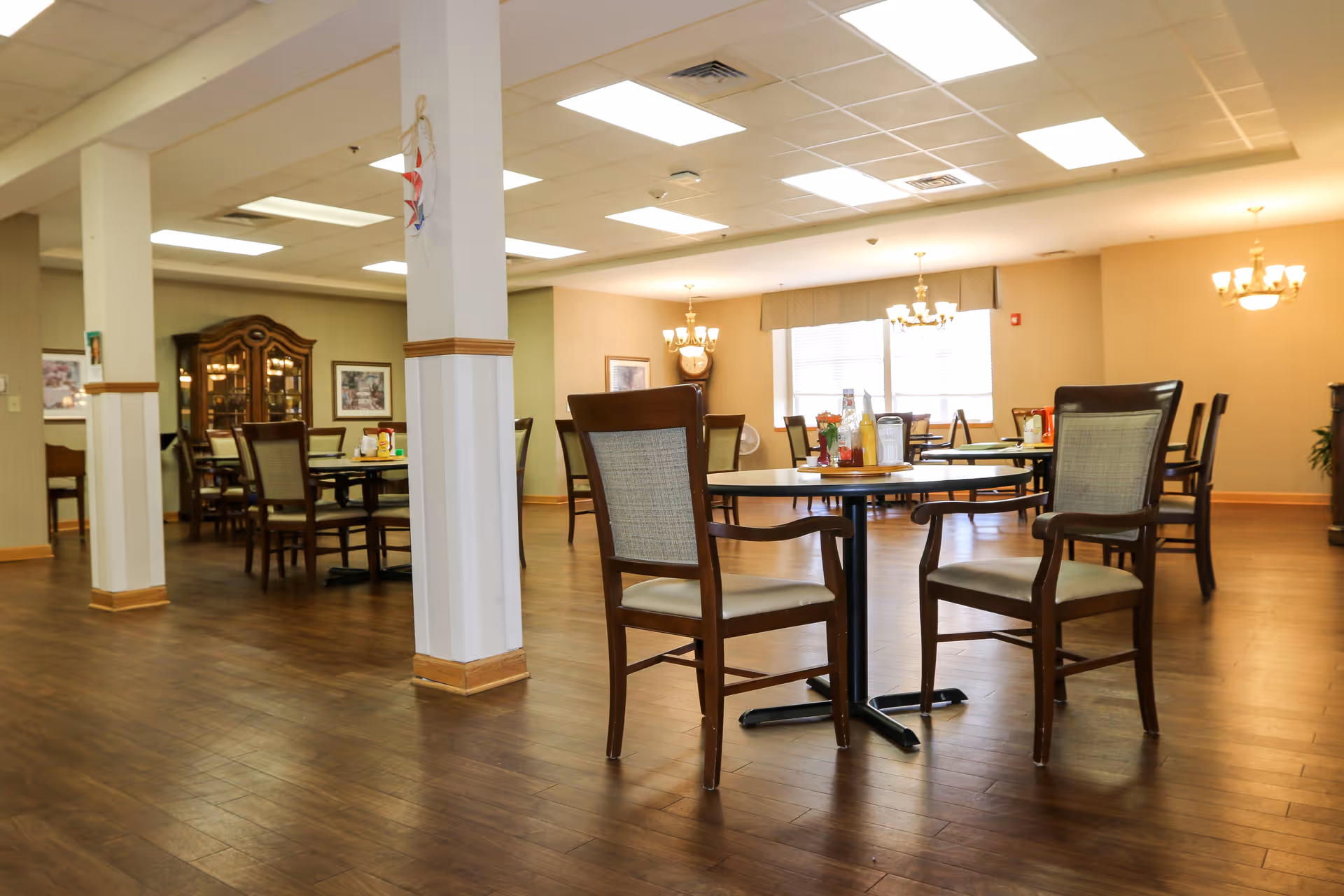 Spacious dining room with round tables and wooden chairs in a senior living facility.