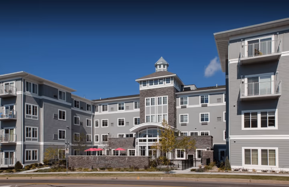 Exterior view of a modern senior living facility named The Waters on 50th, featuring a multi-story building with gray siding, white trim, balconies, and a central entrance with large windows and a small tower on top. There are pink umbrellas on a patio area in front of the building and a clear blue sky above.