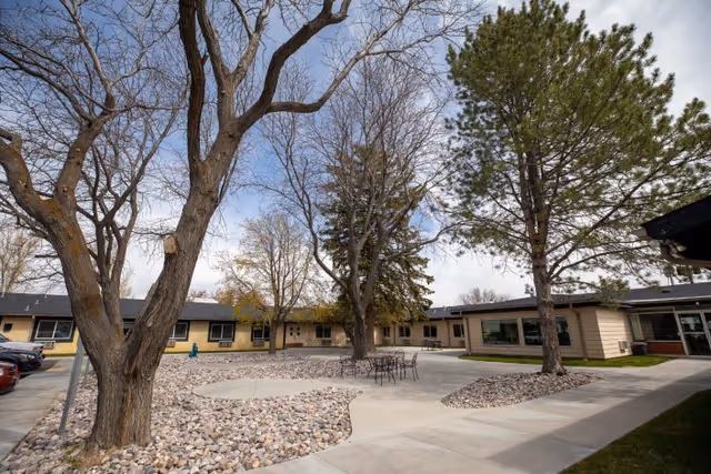 Outdoor courtyard area at Monte Vista Hills Health Care Center with leafless and evergreen trees, a circular concrete patio with metal tables and chairs, surrounded by a building with beige siding and multiple windows under a partly cloudy sky.
