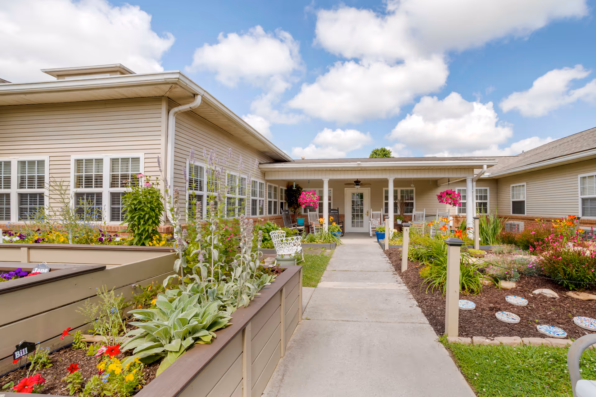 Outdoor garden area at a senior living facility with raised flower beds filled with various plants and flowers. A concrete pathway leads to a covered porch with rocking chairs and hanging flower baskets. The building has beige siding and multiple windows under a partly cloudy sky.