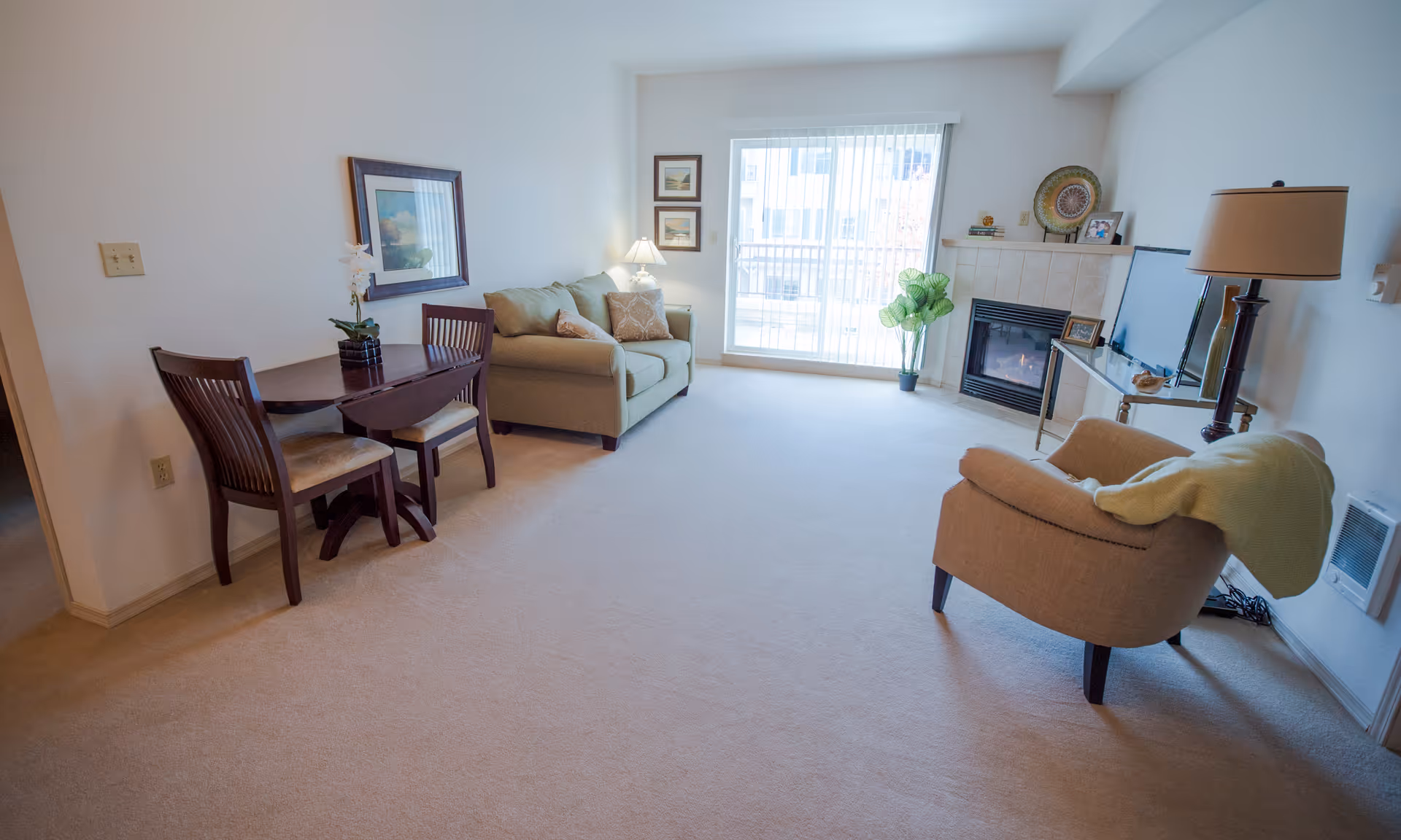 A bright living room with beige carpet, a small wooden dining table with two chairs, a green loveseat with pillows, a beige armchair with a green blanket draped over it, a fireplace with a tiled surround, a flat-screen TV on a glass stand, a floor lamp, framed pictures on the walls, and a sliding glass door with vertical blinds leading to a balcony.