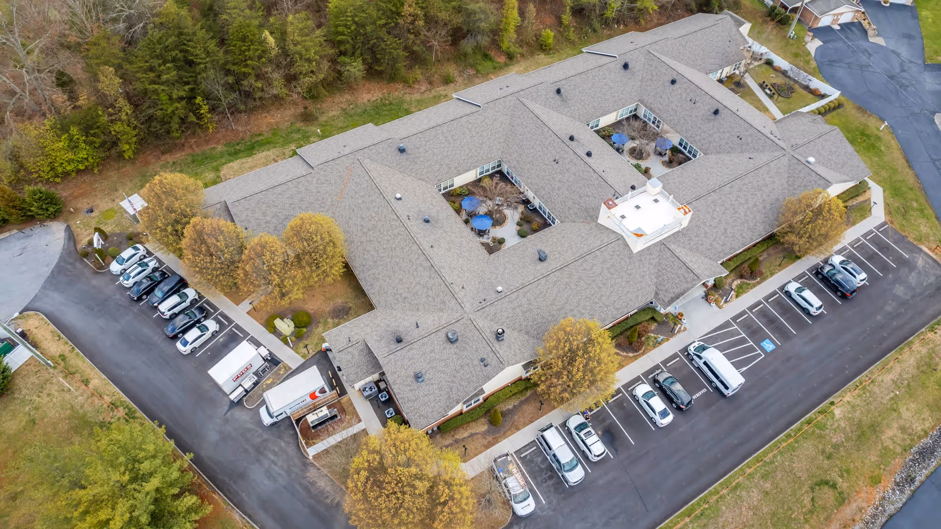 Aerial view of a single-story senior living facility building with a gray roof, surrounded by trees and parking lots with several parked cars. The building has multiple courtyards with outdoor seating areas featuring blue umbrellas.
