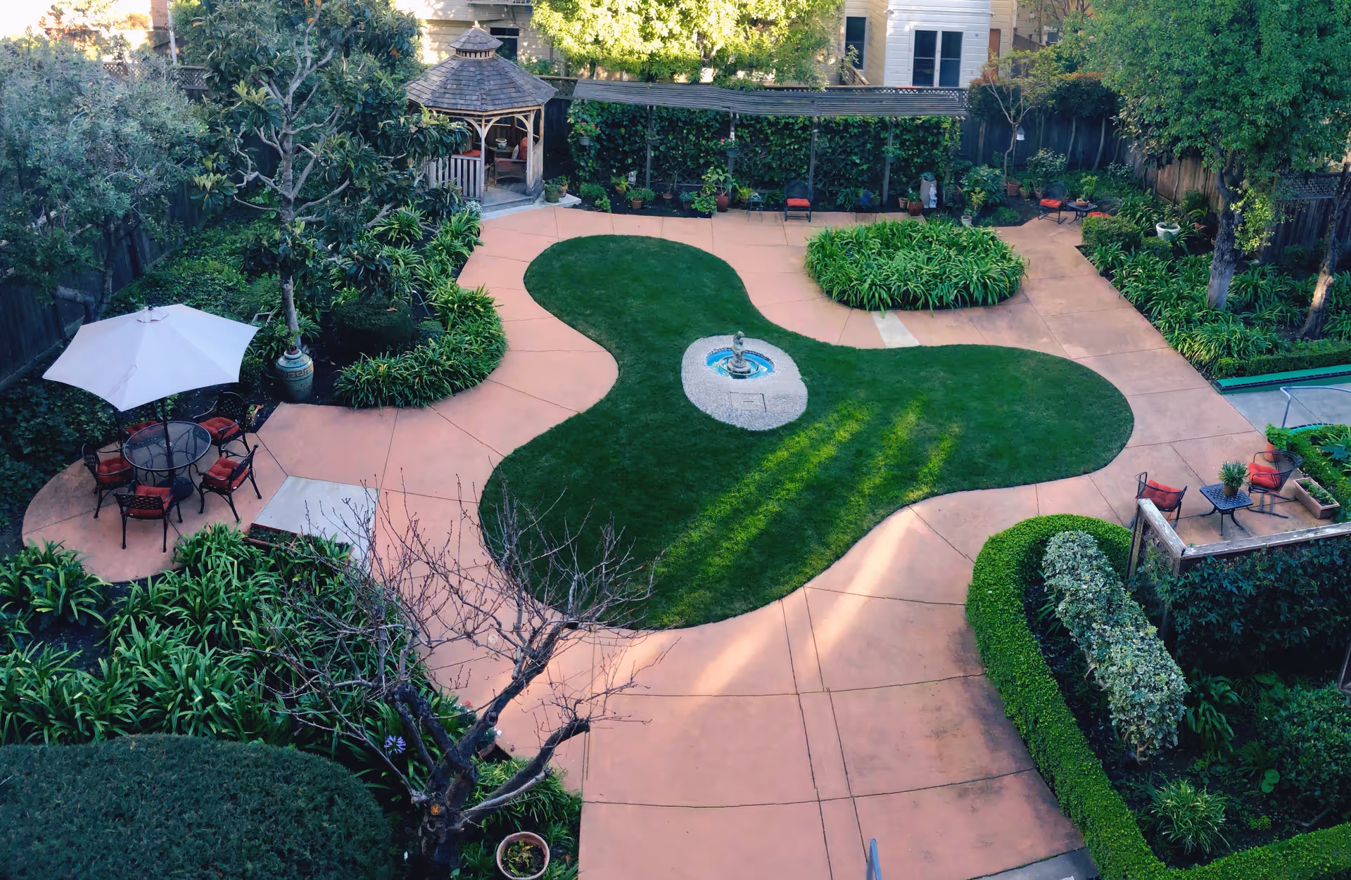 Landscaped courtyard with a kidney-shaped lawn and central fountain, surrounding patio seating, umbrella and a gazebo in the background.