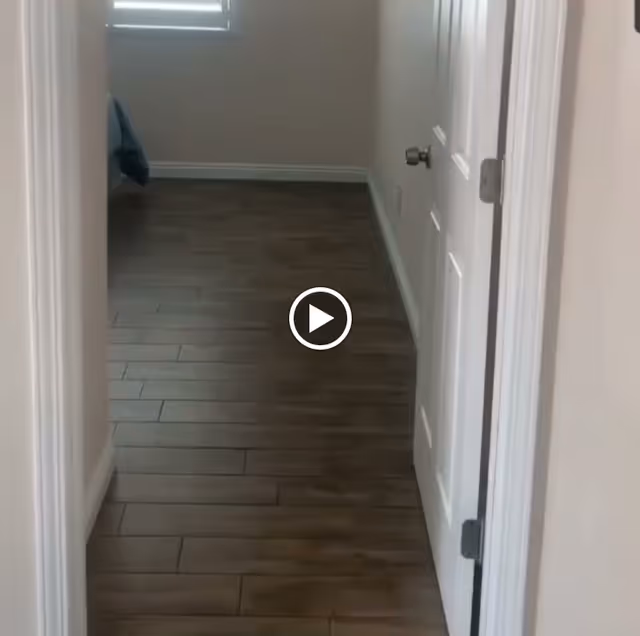 View down a hallway with wood-look tile flooring, white walls, and a white door on the right side. A window with blinds is visible at the end of the hallway.