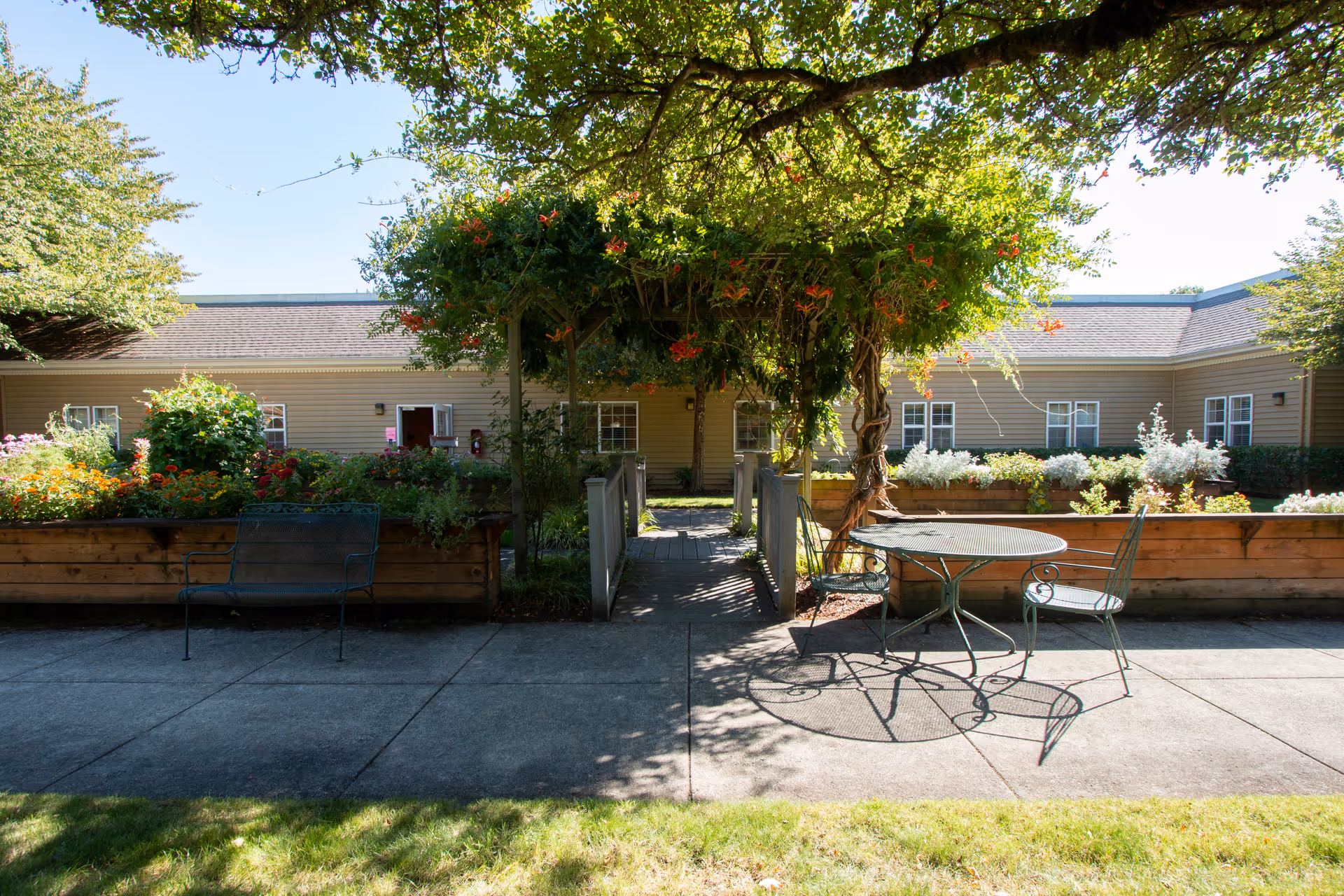 Outdoor courtyard area with raised garden beds filled with various plants and flowers. There is a metal bench on the left and a round metal table with two chairs on the right. A wooden pergola covered with green vines and orange flowers is in the center, providing shade. The courtyard is surrounded by a single-story building with beige siding and multiple windows.