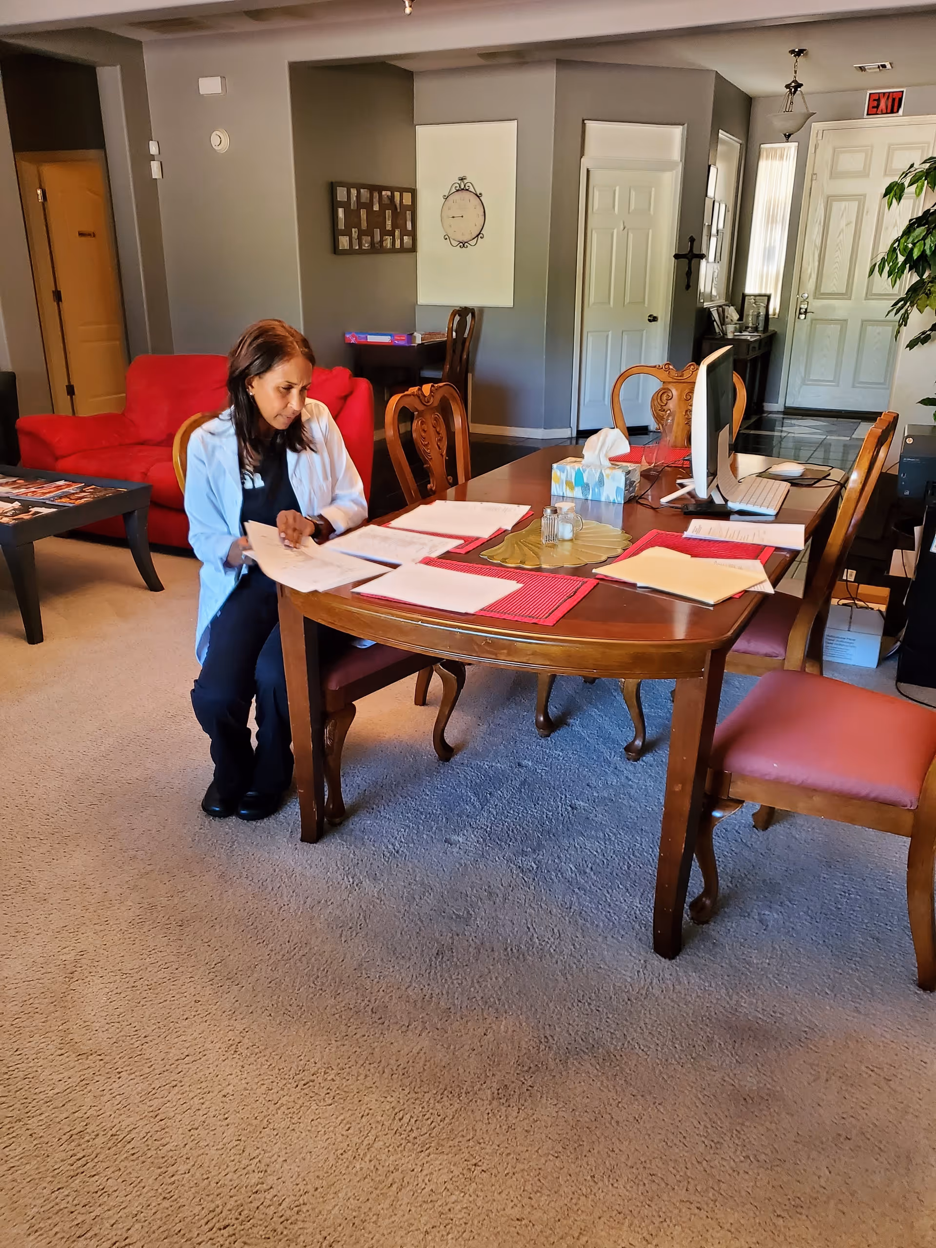 A woman in a white coat sitting on a chair at a wooden dining table covered with papers, a tissue box, and a computer monitor in a well-lit room with beige carpet. In the background, there is a red couch, a small table with chairs, and a white door with an exit sign above it.