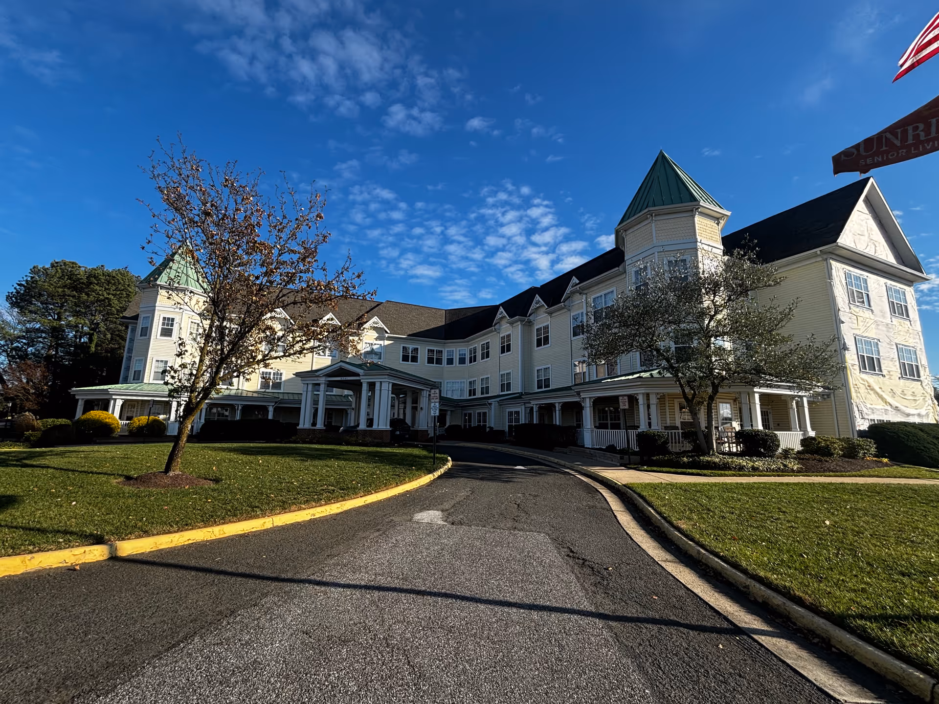 Front exterior of a large three-story yellow senior living building with a circular driveway, lawn, and clear blue sky.