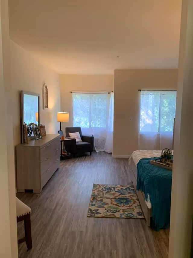 A bright and cozy bedroom with light wood flooring, a bed with a teal blanket and a tray with plants on it, a colorful floral rug, a dresser with framed photos and a lamp, a comfortable armchair with a pillow, and two large windows with sheer white curtains letting in natural light.