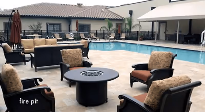 Outdoor patio area with cushioned chairs arranged around a circular fire pit, a swimming pool in the background, and additional seating with umbrellas near a building with a tiled roof.
