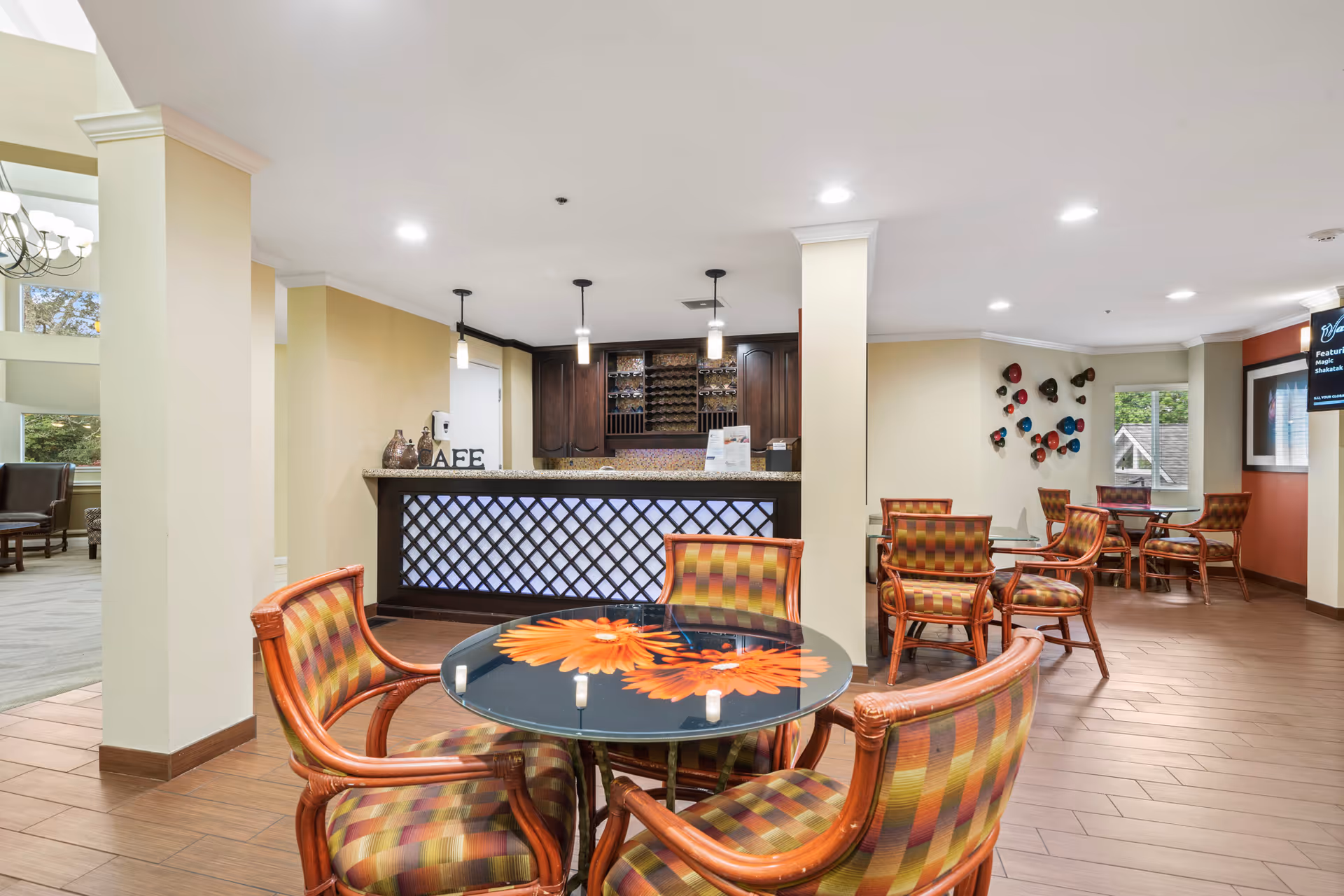 Interior view of a senior living facility lounge area with multiple round tables and cushioned chairs featuring a checkered pattern. A counter with a lattice design and pendant lights hangs above it. The walls are painted in warm tones, and there is decorative wall art and a window letting in natural light.