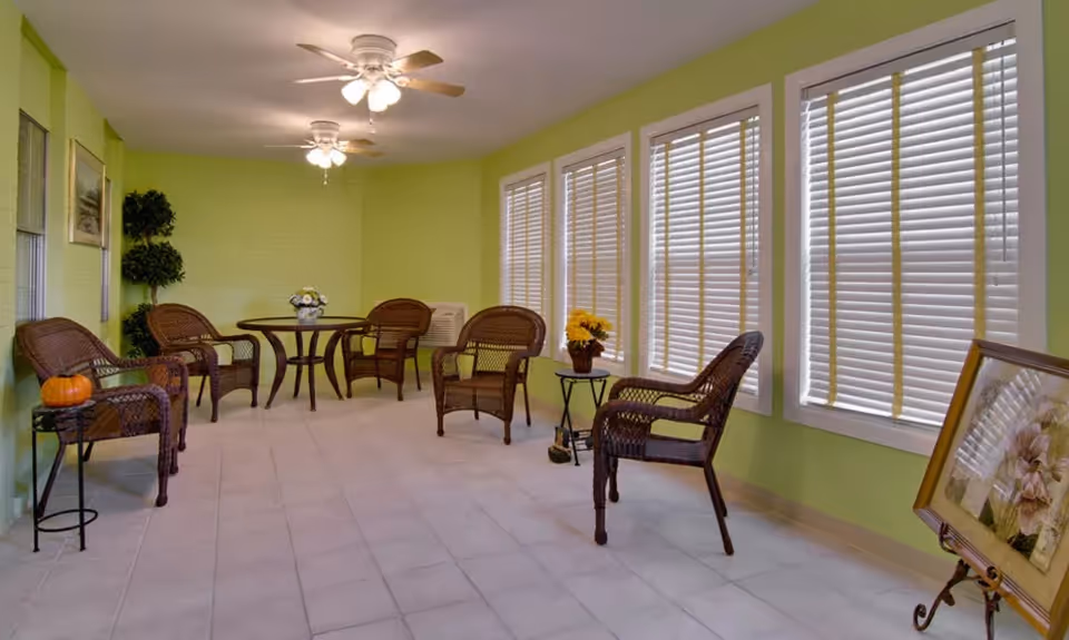 Sunroom-style communal seating area with wicker chairs and tables against light green walls and large shuttered windows.