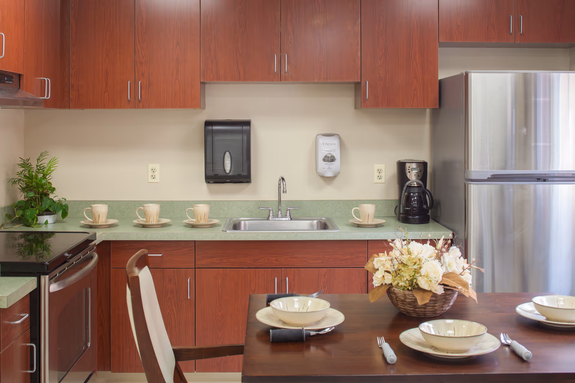 A kitchen area with wooden cabinets, a stainless steel refrigerator, a stove, a sink, and a coffee maker. The countertop has several cups and saucers, and a dining table in the foreground is set with bowls, plates, forks, and a floral centerpiece.