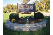 Outdoor view of a stone-bordered garden bed with a sign that reads 'Apple Rehab Saybrook' and the address '433 Boston Post Rd' surrounded by green grass and trees in the background.