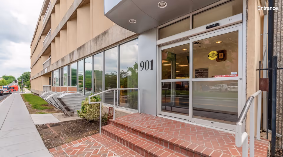 Entrance of a building with the number 901 displayed on a metal panel next to glass automatic sliding doors. There are brick steps leading up to the entrance with metal handrails on both sides. The building has large windows along the side and a sidewalk runs parallel to it. A yellow caution sign is visible on the door.