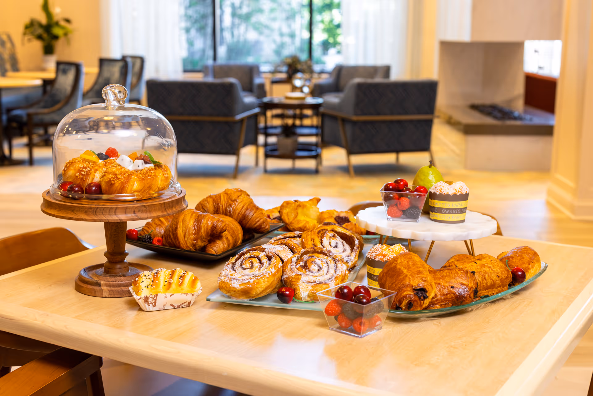 A wooden table in a cozy living area with an assortment of pastries including croissants, cinnamon rolls, and muffins, along with small cups of berries and a pear. In the background, there are comfortable armchairs and a fireplace near large windows with sheer curtains.