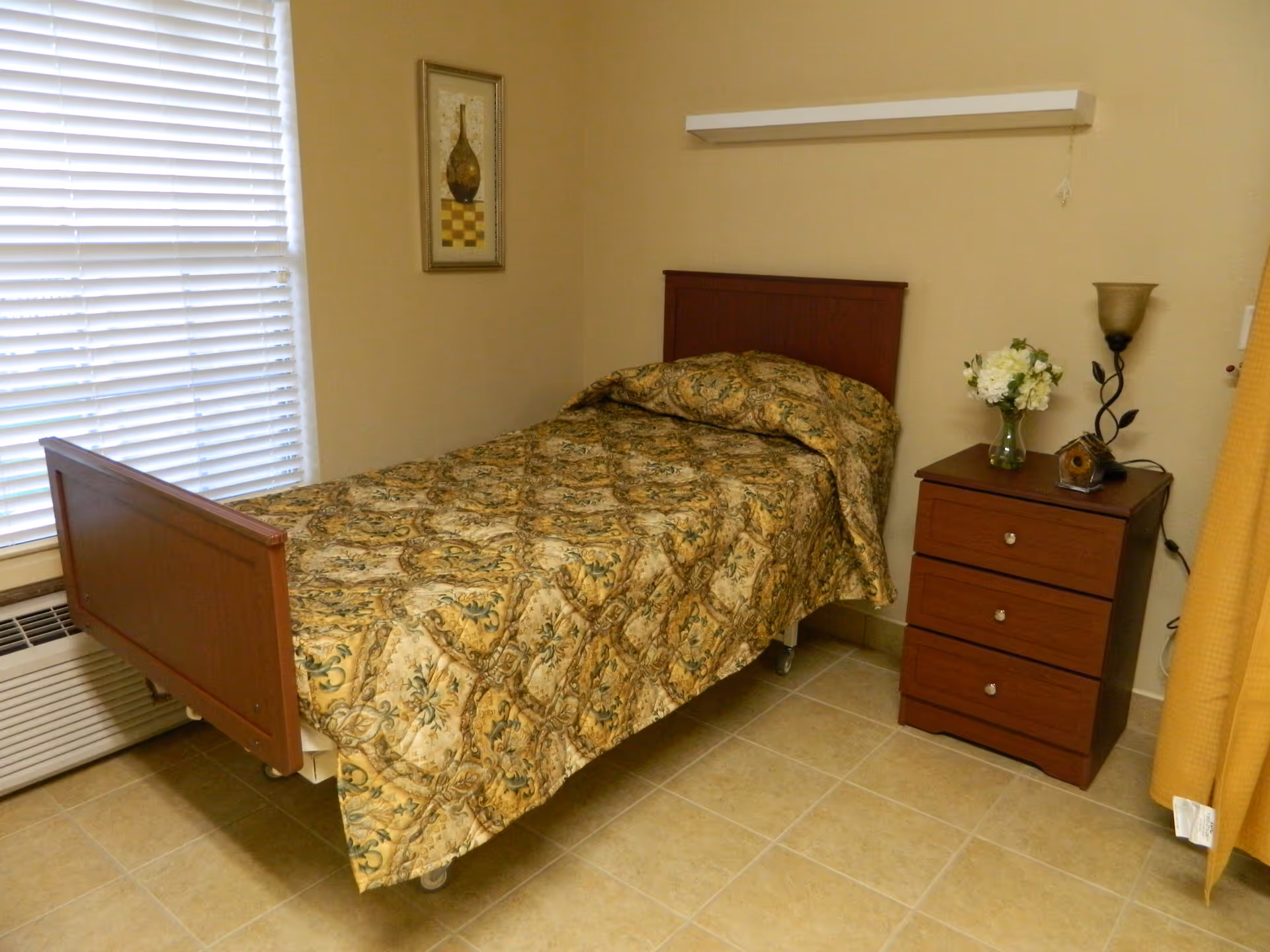 A small bedroom with a single bed covered in a patterned quilt. Next to the bed is a wooden nightstand with three drawers, a vase with white flowers, a small decorative birdhouse, and a lamp. There is a window with closed blinds on the left side and a framed picture on the wall above the bed.