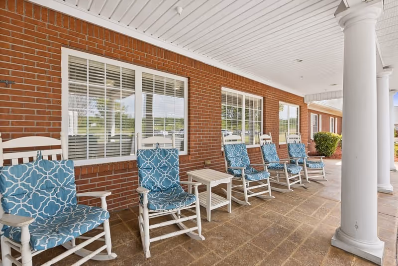 Covered outdoor patio area with a row of white rocking chairs featuring blue patterned cushions, positioned in front of a brick wall with large windows and white trim. The patio has a tiled floor and white columns supporting the roof.