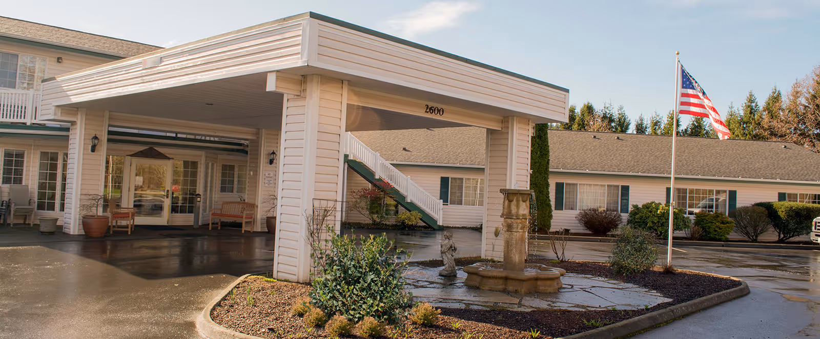Exterior view of a senior living facility with a covered entrance, benches, potted plants, a small fountain, and an American flag on a flagpole. The building is light-colored with green shutters and surrounded by trees and shrubs.
