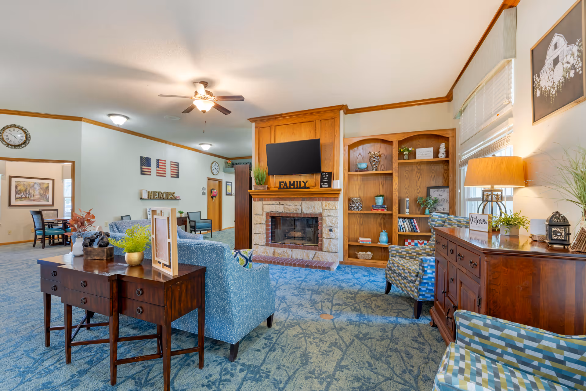 A cozy living room area in a senior living facility featuring a stone fireplace with a mounted TV above it. The room has blue patterned carpet, wooden bookshelves with decorative items, comfortable armchairs with patterned upholstery, a wooden sideboard with a lamp and plants, and a wooden table with plants and decorative pieces. The walls are light-colored with crown molding, and there are windows with blinds letting in natural light. In the background, there is a dining area with tables and chairs, and wall decorations including clocks and American flag art.