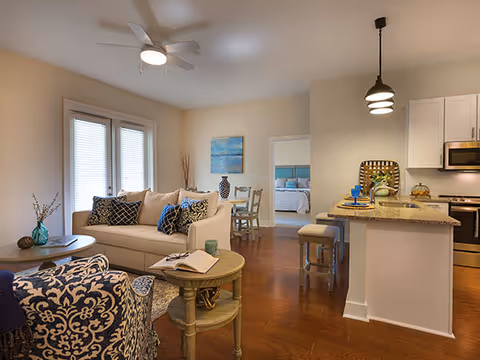 A cozy living room with a beige sofa adorned with patterned cushions, a round wooden coffee table with an open book, and a blue patterned armchair. The room has wooden flooring, a ceiling fan with light, and a kitchen area with a granite countertop and bar stools. In the background, a bedroom with a bed and blue bedding is visible through an open doorway.