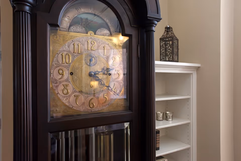 Close-up view of a dark wooden grandfather clock with a decorative clock face showing the time as 4:22. In the background, there is a white shelving unit with a decorative lantern on top and two small silver candle holders on one of the shelves.