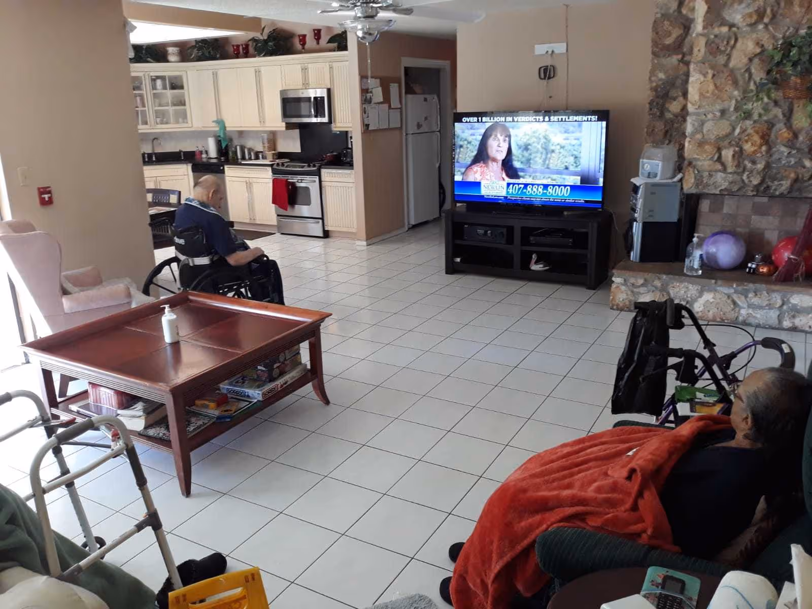 A living room area in an assisted living facility with two elderly individuals, one in a wheelchair and the other seated in a recliner covered with a red blanket. The room features a tiled floor, a wooden coffee table with a bottle of hand sanitizer on top, a TV displaying a commercial, a stone fireplace, and a kitchen area visible in the background.