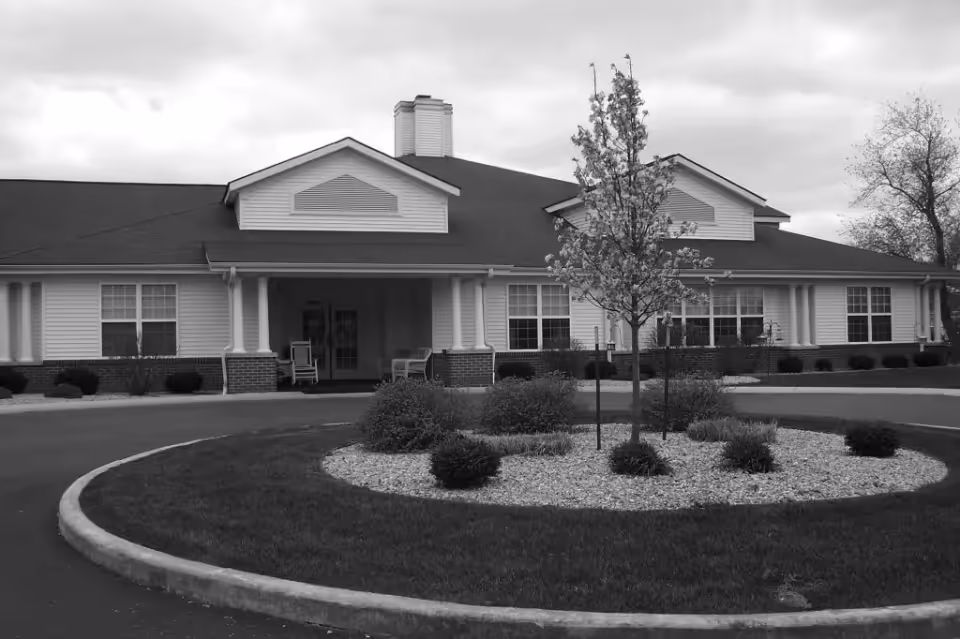 Front exterior view of a single-story building with a covered entrance, two rocking chairs on the porch, a circular driveway with a landscaped island featuring small bushes and a young tree, under a cloudy sky.