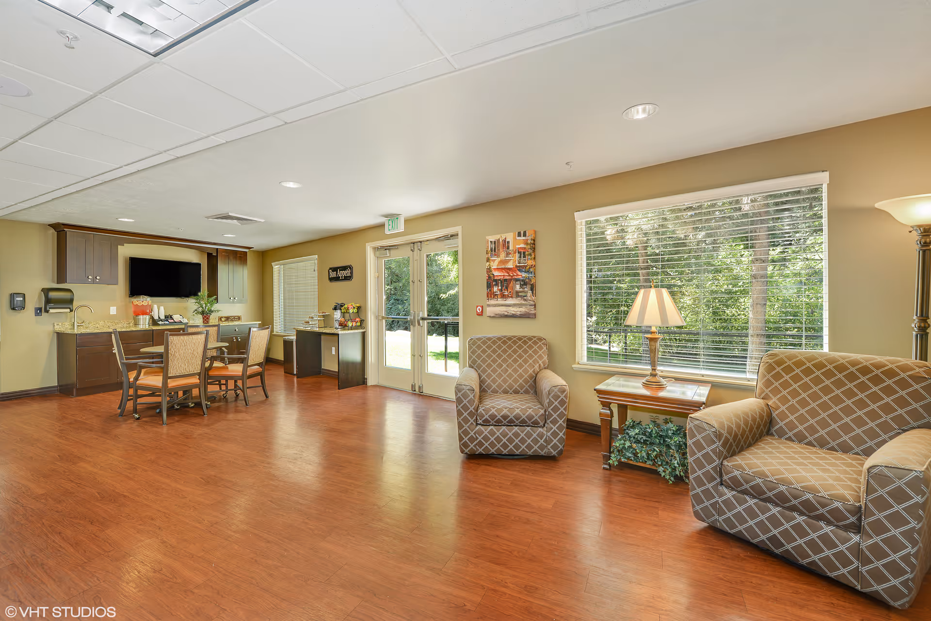 A bright and spacious common area in a senior living facility featuring a small dining table with four chairs, a kitchenette with dark cabinets, a wall-mounted TV, and a countertop with snacks. There are two patterned armchairs and a matching loveseat near a large window with blinds, a side table with a lamp, and a floor lamp. Double glass doors lead outside, and the room has wood flooring and beige walls.