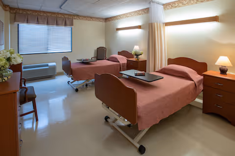 A clean and well-lit room with two hospital-style beds covered in pink bedspreads. Each bed has an overbed table, and there is a wooden nightstand with a lamp between the beds. A window with closed blinds and a valance is on the left wall, along with a chair and a small table with flowers. The room has light-colored walls and flooring, with a privacy curtain partially drawn around one bed.