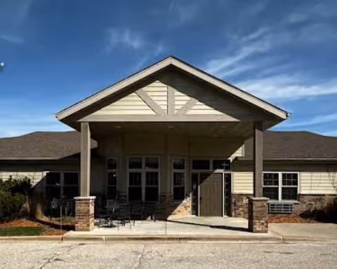Front exterior view of a single-story building with a covered entrance supported by two columns with brick bases, outdoor seating, and a clear blue sky above.