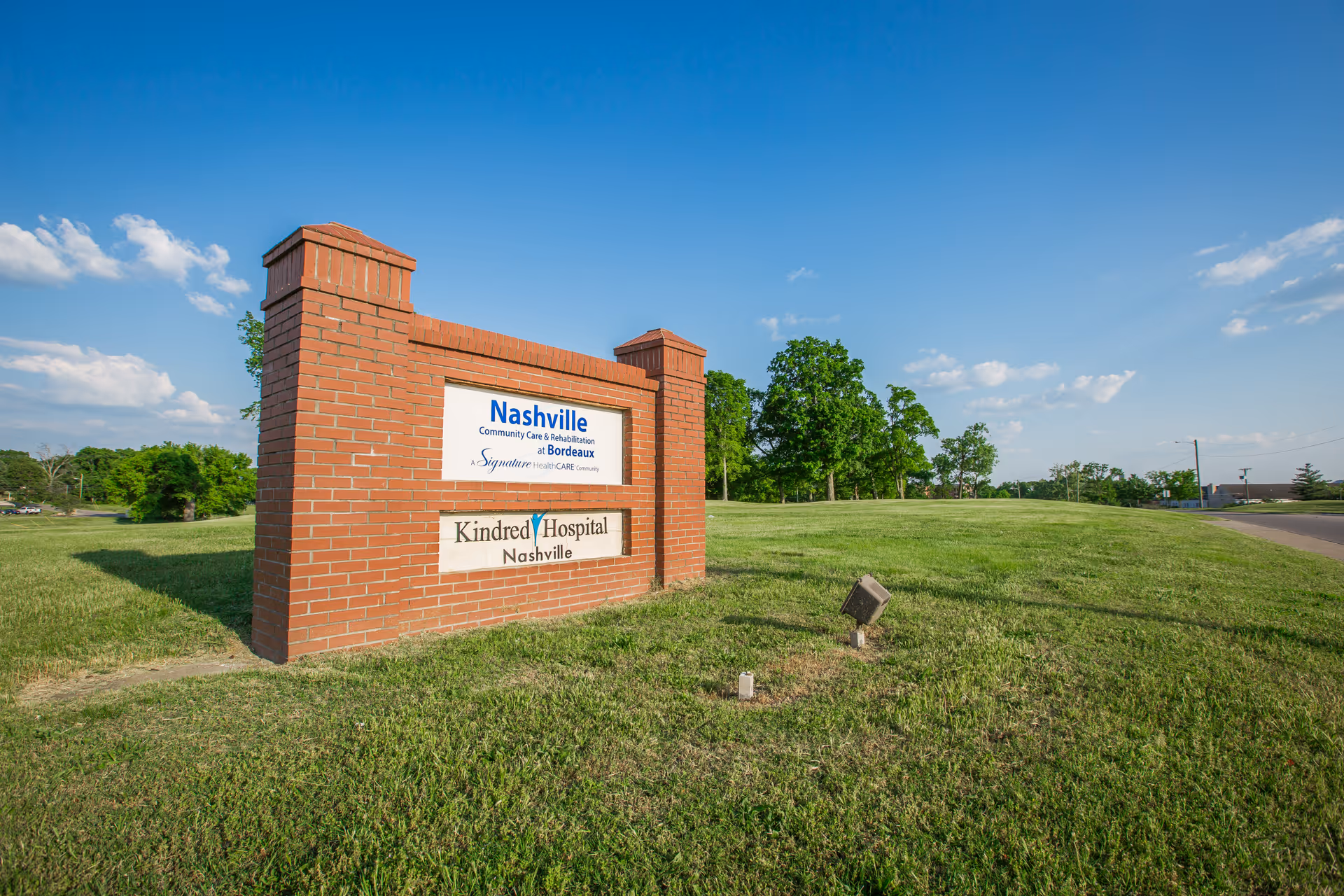 A brick sign on a grassy area under a blue sky with some clouds. The sign reads 'Nashville Community Care & Rehabilitation at Bordeaux' and 'Kindred Hospital Nashville'. Trees and a road are visible in the background.