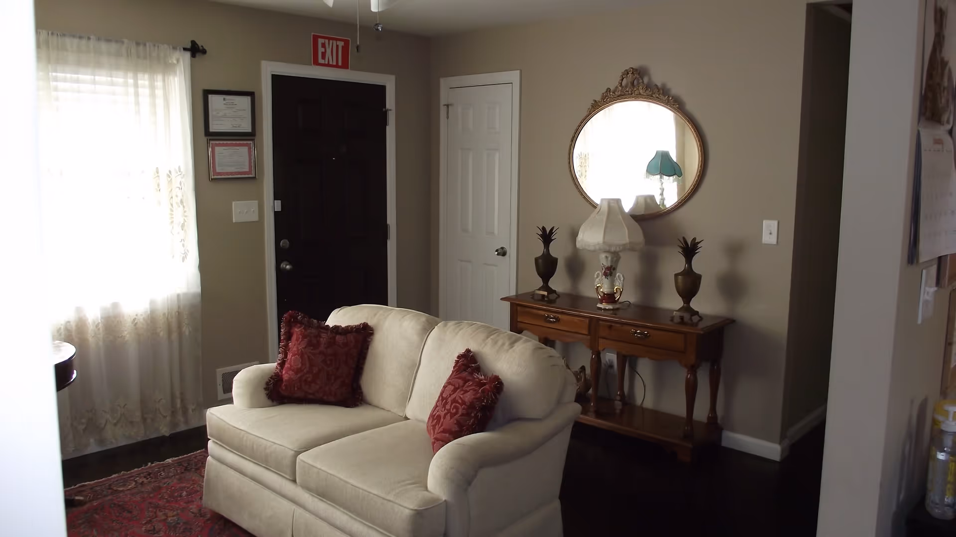 A cozy living room area with a cream-colored loveseat adorned with two red decorative pillows. Behind the loveseat is a wooden console table with two small decorative plants and a lamp with a white shade. Above the table hangs an ornate oval mirror reflecting a window and a blue lamp. To the left, there is a window with sheer curtains allowing natural light to enter. A dark front door with an exit sign above it and a white door are visible in the background.