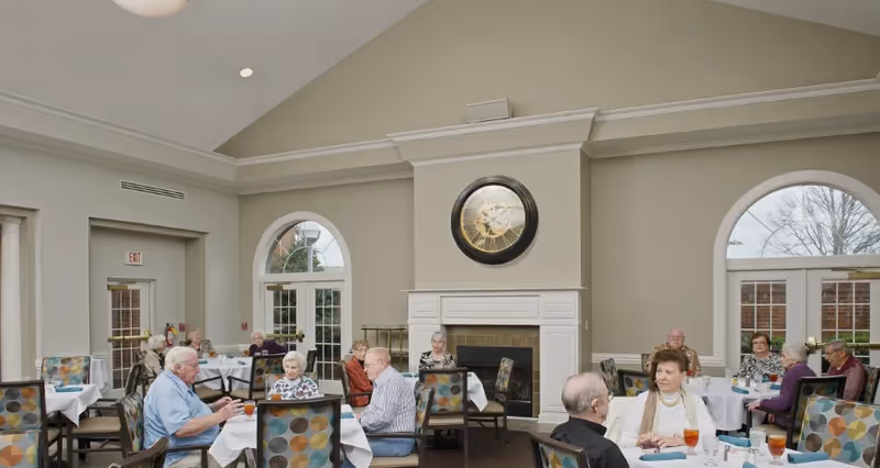 Seniors sitting at round dining tables in a bright common dining room with a fireplace and large wall clock.