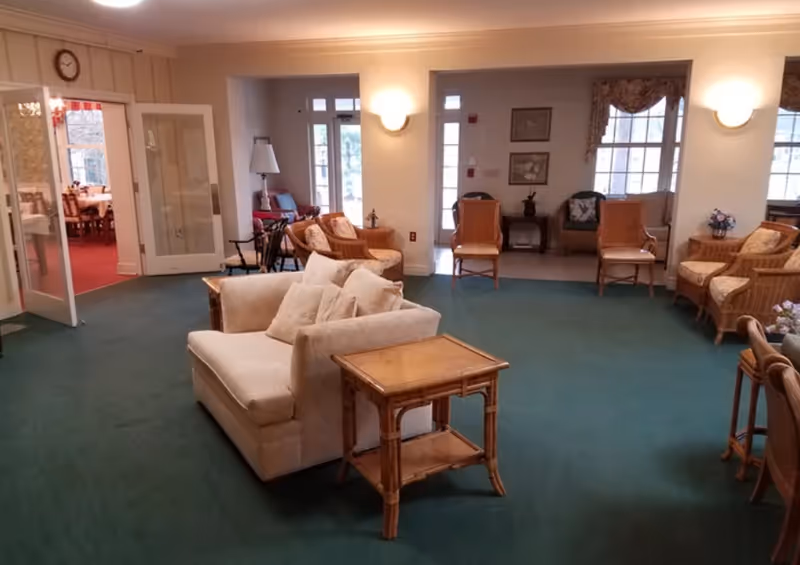 A cozy living room area with a green carpet, beige loveseat with cushions, wooden side table, and several wicker chairs arranged around the room. The room is well-lit with wall sconces and natural light coming through large windows. In the background, a dining room with a red carpet and wooden dining chairs is visible through open double doors.