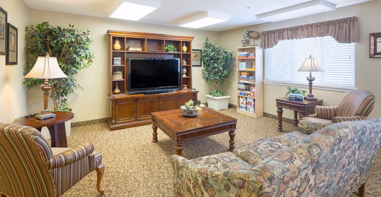 Communal living room with patterned sofas and armchairs arranged around a wooden coffee table and television cabinet, with lamps, plants, and a bookshelf by a window.