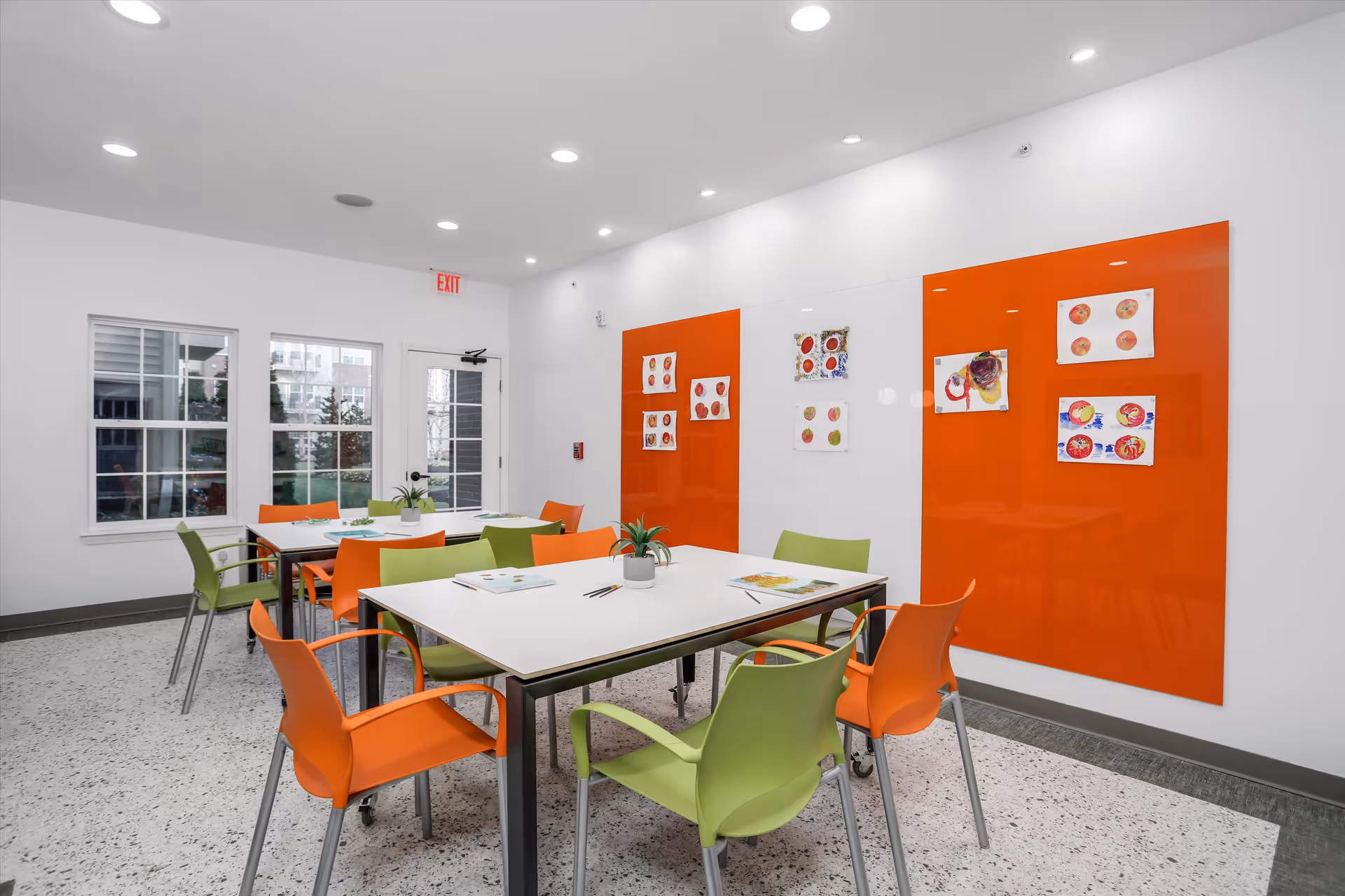A bright, modern activity room with white walls and ceiling, featuring two white tables surrounded by orange and green chairs. The walls have large orange and white panels with children's artwork displayed. There are windows and a glass door letting in natural light, and small potted plants are placed on the tables.