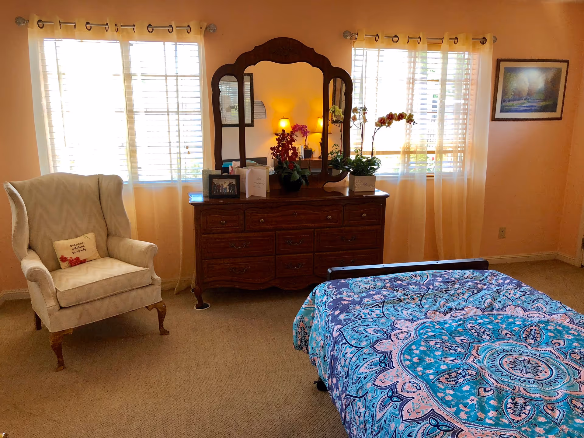 A cozy bedroom with a patterned blue and pink bedspread on a bed in the foreground. There is a wooden dresser with a large ornate mirror and decorative plants on top, positioned between two windows with sheer curtains. To the left, there is a beige upholstered armchair with a small pillow. The walls are painted a warm peach color, and a framed picture hangs on the right wall.