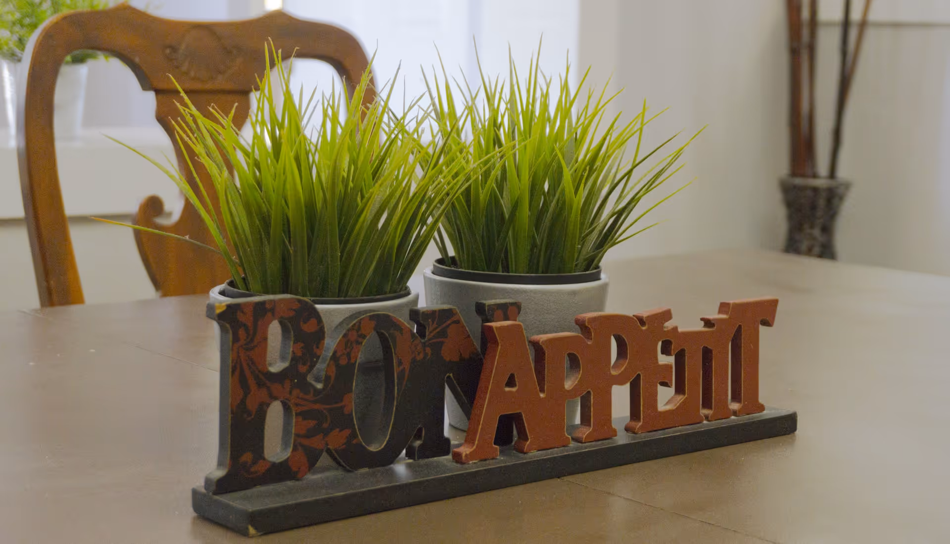 A close-up view of a wooden dining table with two small potted green plants and a decorative sign that reads 'Bon Appétit'. In the background, there is a wooden chair and a window with white curtains.