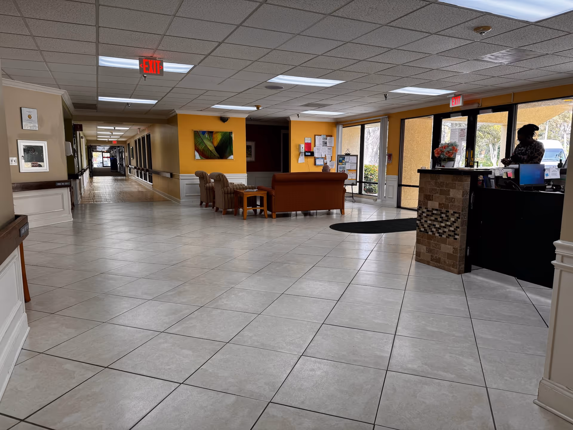 Interior view of a senior living facility lobby with tiled floor, a reception desk on the right with a person standing behind it, seating area with chairs and a sofa in the middle, and a long hallway extending to the left. The walls are painted yellow and beige, and there are large windows near the entrance letting in natural light.