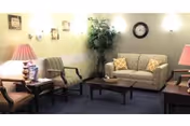 A cozy living room with a beige patterned sofa adorned with two yellow patterned pillows, two armchairs with striped upholstery, a wooden coffee table, side tables with pink lamps, a wall clock, and a potted plant in the corner.