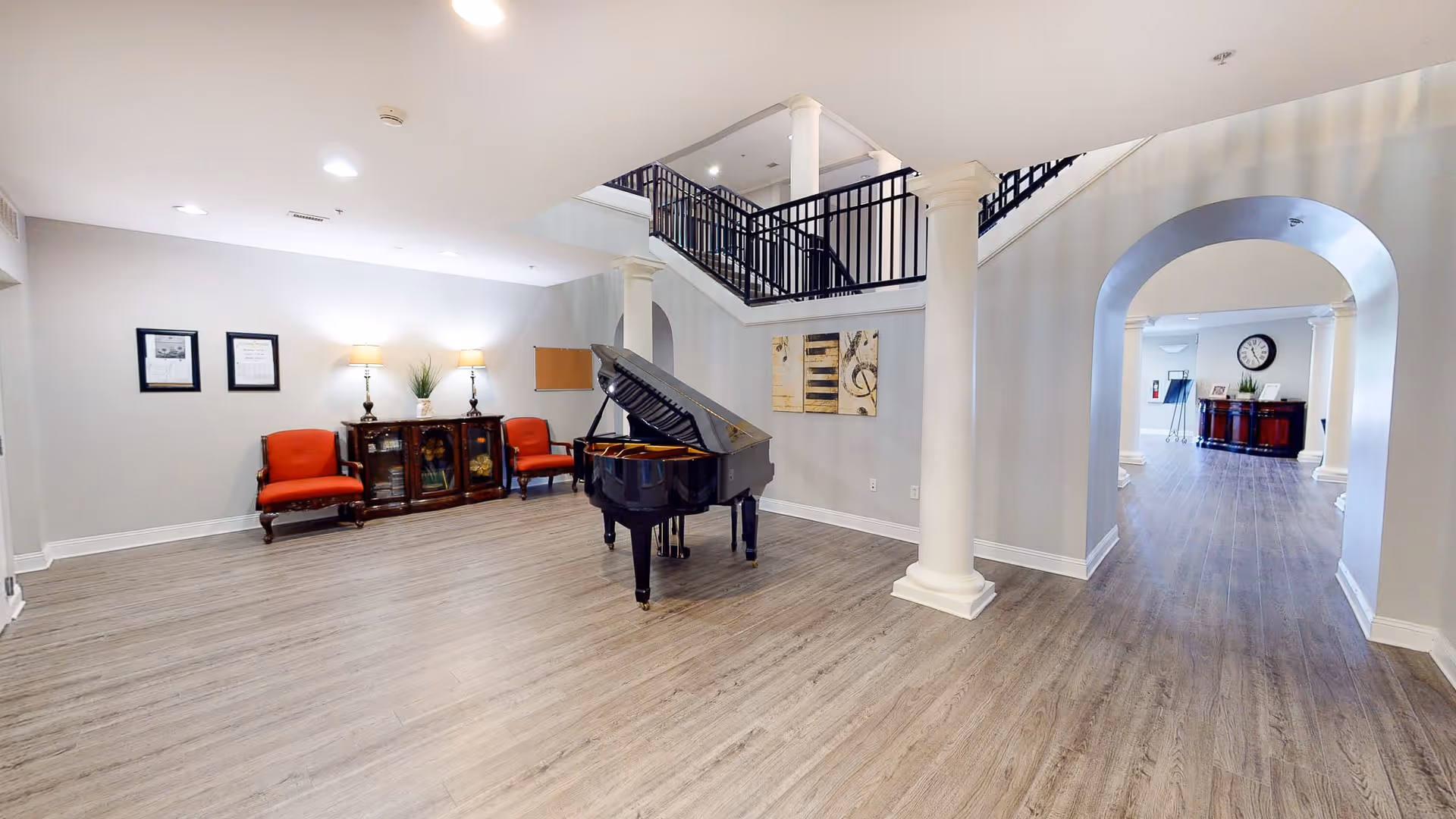 Spacious interior room with a grand piano in the center, two red upholstered chairs and a wooden cabinet against the wall, decorative framed pictures and lamps on the cabinet, white columns, and an open staircase with black railings leading to an upper floor. The room has light-colored walls and wood flooring, with an arched doorway leading to another area with more furniture and a clock on the wall.