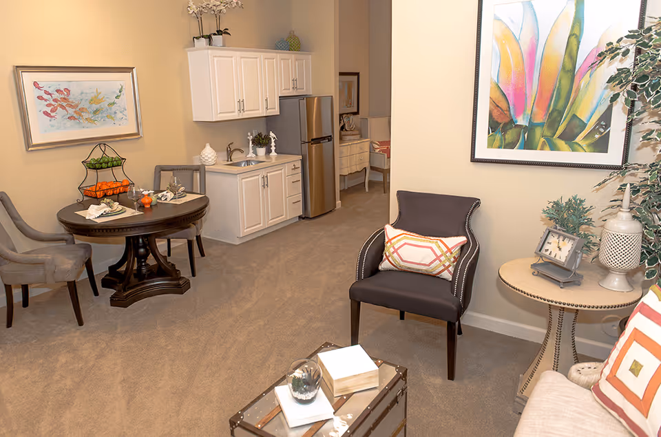 Cozy assisted-living suite with armchairs, a small round dining table, a kitchenette with sink and refrigerator, and colorful wall art.