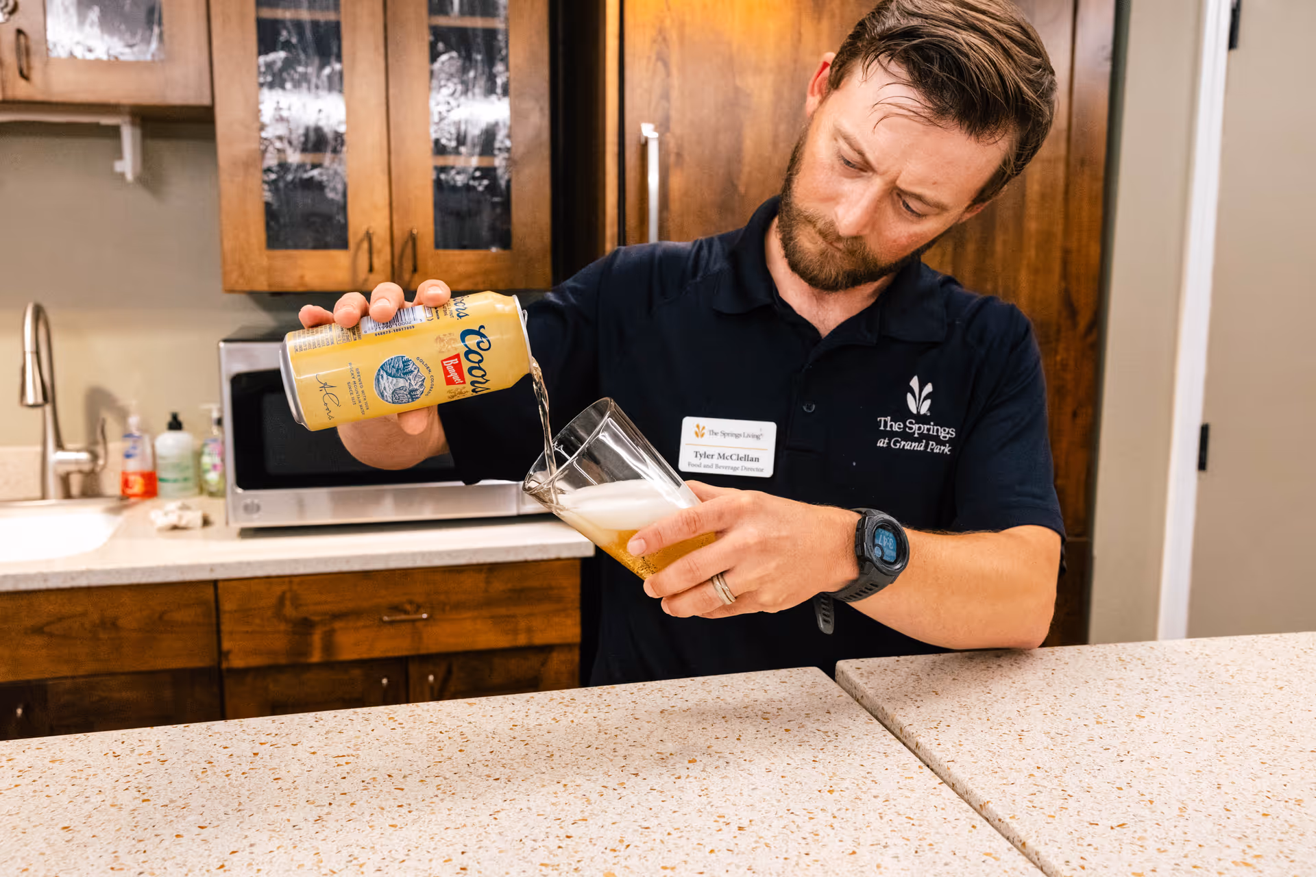 A man wearing a navy blue polo shirt with The Springs at Grand Park logo is pouring a can of Coors beer into a glass in a kitchen area with wooden cabinets, a microwave, and a sink in the background.