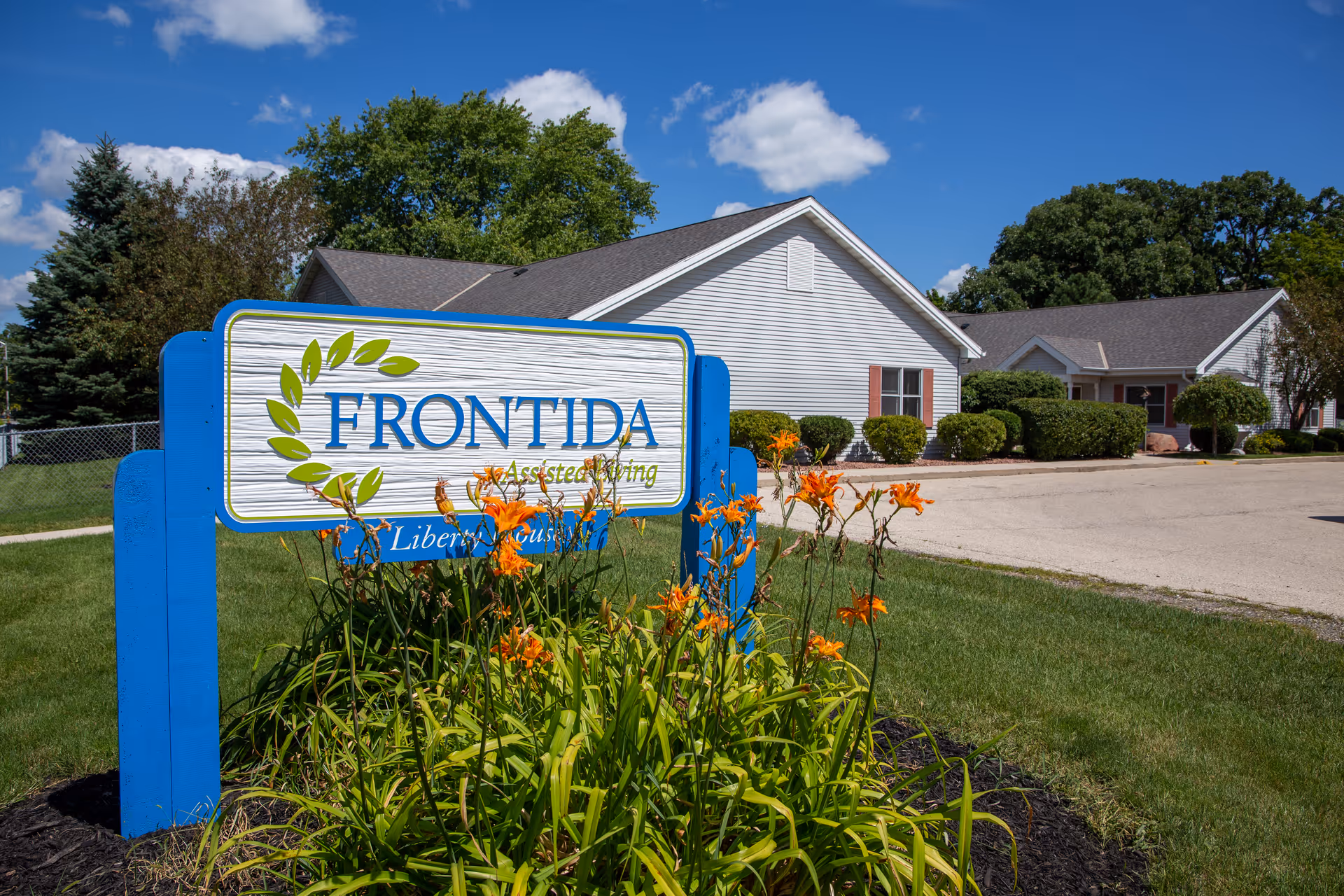 Outdoor view of Frontida Assisted Living: Liberty House facility sign surrounded by orange flowers and green plants, with a white building and blue sky with clouds in the background.