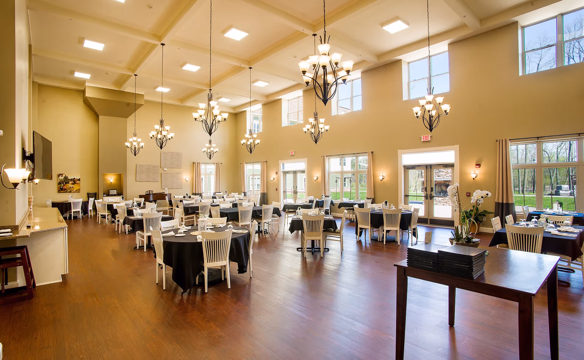Spacious dining room with multiple round and rectangular tables set, high ceilings, chandeliers, and large windows overlooking greenery.