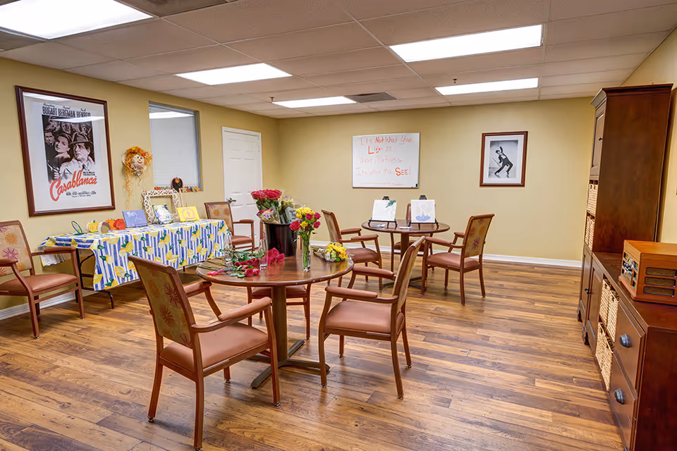 A bright community activity room with round tables and chairs, floral centerpieces and a decorated side table against a yellow wall.