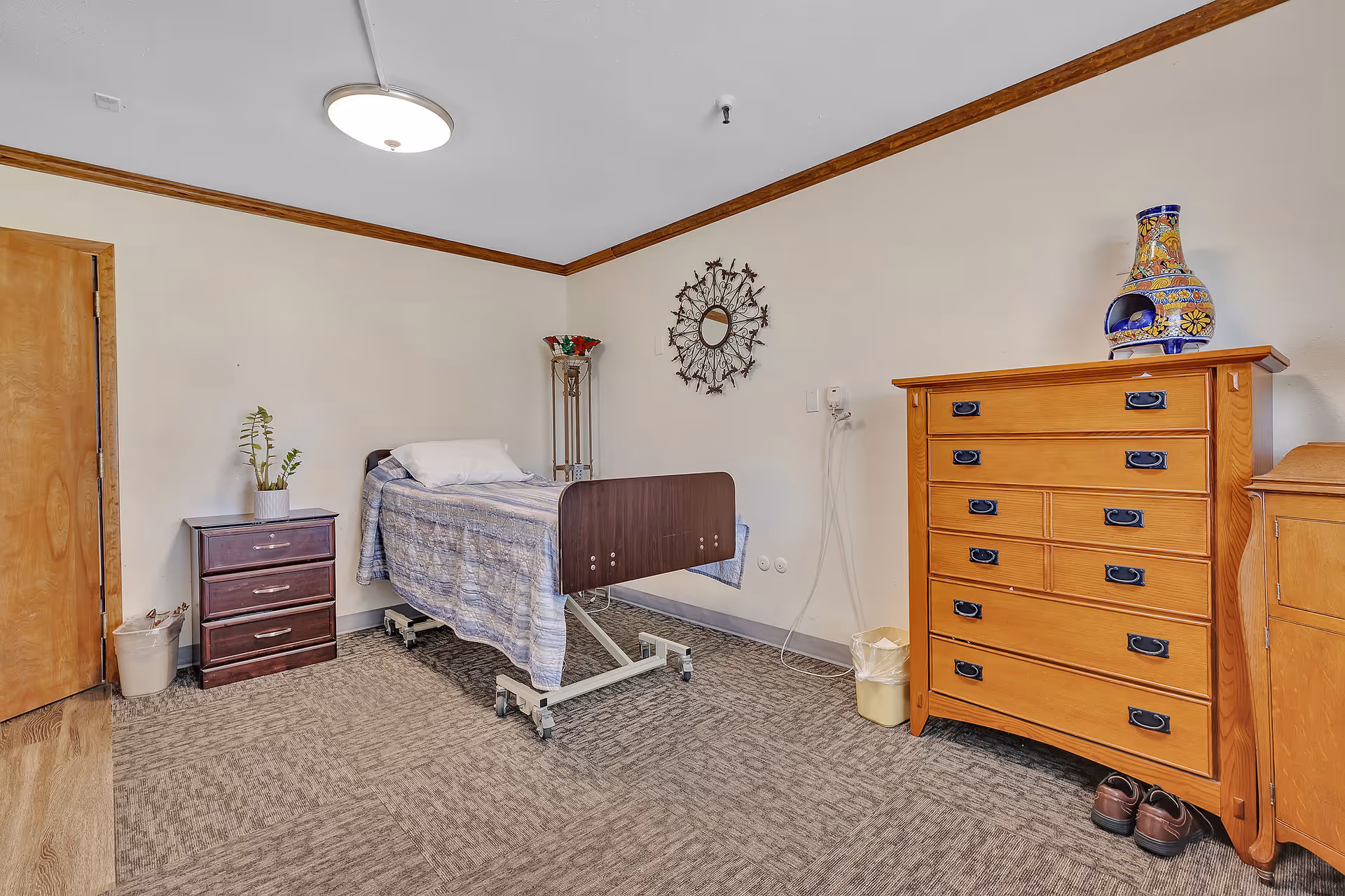 A simple, clean bedroom in a care facility with a single hospital-style bed covered with a blue patterned blanket. The room has beige walls with wooden trim, a wooden door, a small dark wooden nightstand with a plant on top, a tall decorative lamp, a wall-mounted decorative mirror, a wooden dresser with multiple drawers, a colorful ceramic vase on top, and a pair of brown shoes on the floor next to the dresser. The floor is carpeted in a neutral tone.